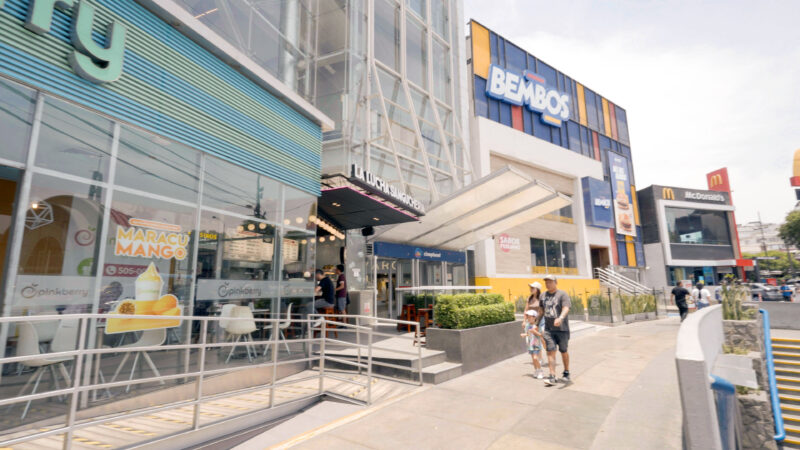 Facha comercial con diversas marcas de retail y restaurantes en avenida urbana con alto tránsito peatonal.